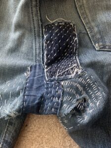 Several patches made into the crotch of a pair of blue jeans made with blue denim scraps and white thread