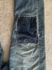 Close up of a rectangular piece of dark blue denim used to patch the knee of a pair of blue jeans. The patch has been embroidered with white thread in a geometric pattern