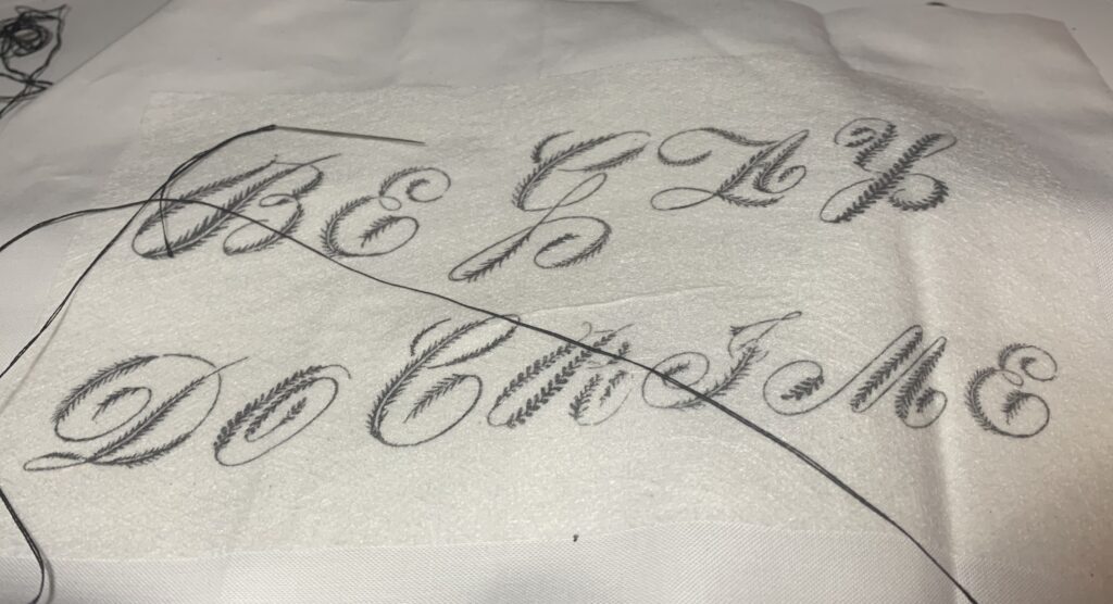 A piece of white cotton with an embroidery pattern reading "Be Gay Do Crime" in a florid script